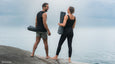 A man and woman stand barefoot on a rocky shore, holding Manduka yoga mats, gazing at a calm ocean under a cloudy sky.