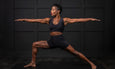 A woman is demonstrating a warrior yoga pose on a black mat against a dark wall, showcasing strength and balance.