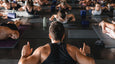 A yoga instructor leads a group yoga class, with participants in various poses on mats in a spacious, well-lit studio.