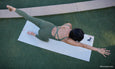 A person performs a yoga pose on a mat in an outdoor setting, wearing a fitted olive green outfit, with one arm extended forward.