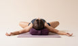 A person lies on a yoga mat with arms extended, resting their head on a purple pillow in a serene, light-colored environment.