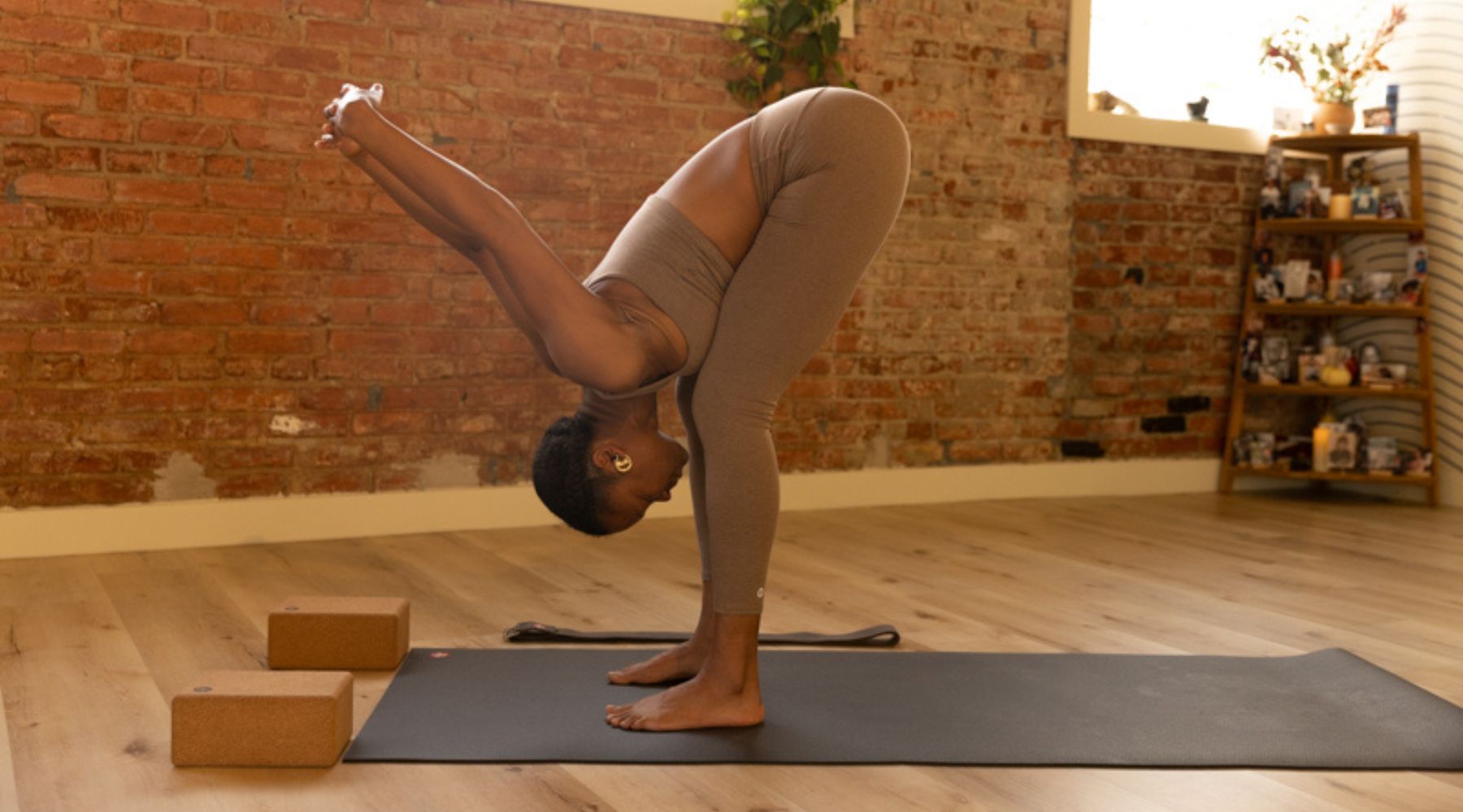A woman in a fitted brown outfit performs a forward bend yoga pose in a cozy, brick-walled studio with wooden decor.