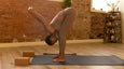 A woman in a fitted brown outfit performs a forward bend yoga pose in a cozy, brick-walled studio with wooden decor.