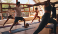 A group of individuals practicing hot yoga in a spacious, sunlit studio with large windows and wooden floors.