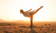 Silhouetted yoga teacher striking a high kick against a glowing sunset, surrounded by grassy fields and distant mountains.